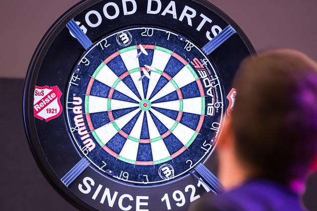 Ein Spieler wirft einen Pfeil auf ein beleuchtetes Dartboard mit dem Schriftzug „GOOD DARTS“ im Hintergrund.