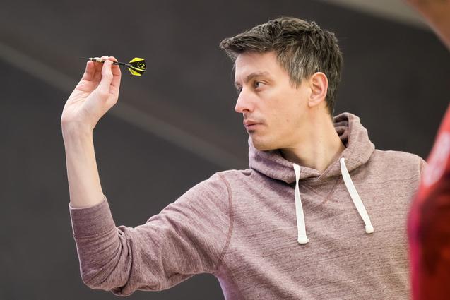 Ein Mann in einem grauen Hoodie zielt mit einem Dart auf eine Dartscheibe in einer Turnierumgebung.