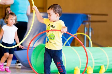 Zwei Kinder spielen in einer Turnhalle mit hula-hoops und einem grünen Tunnel. Ein Kind hält zwei Reifen in der Hand.