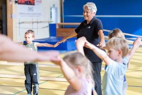 Seniorin leitet Kinder in einer Sporthalle an, während sie ihre Arme in die Höhe strecken.