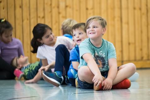 Gruppe von Kindern sitzt in einer Turnhalle, einige lachen, während ein Junge mit roten Schuhen lächelt.